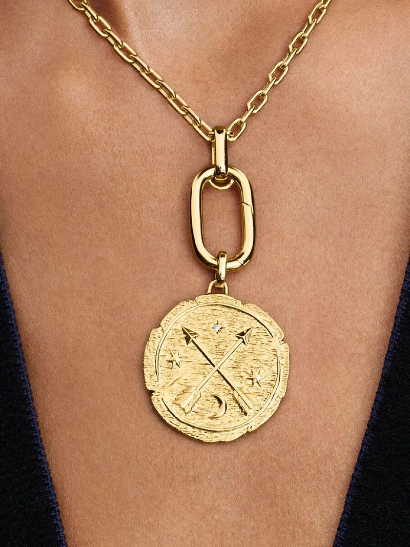 Medaljong Korsande Pilar Berlock Guld. Symbol för att sträva mot det bättre. Tillverkad av 14 karat guldplätering med korsade pilar och latinska frasen "AD MELIORA". En påminnelse om att följa din inre kompass och leva efter dina värderingar.